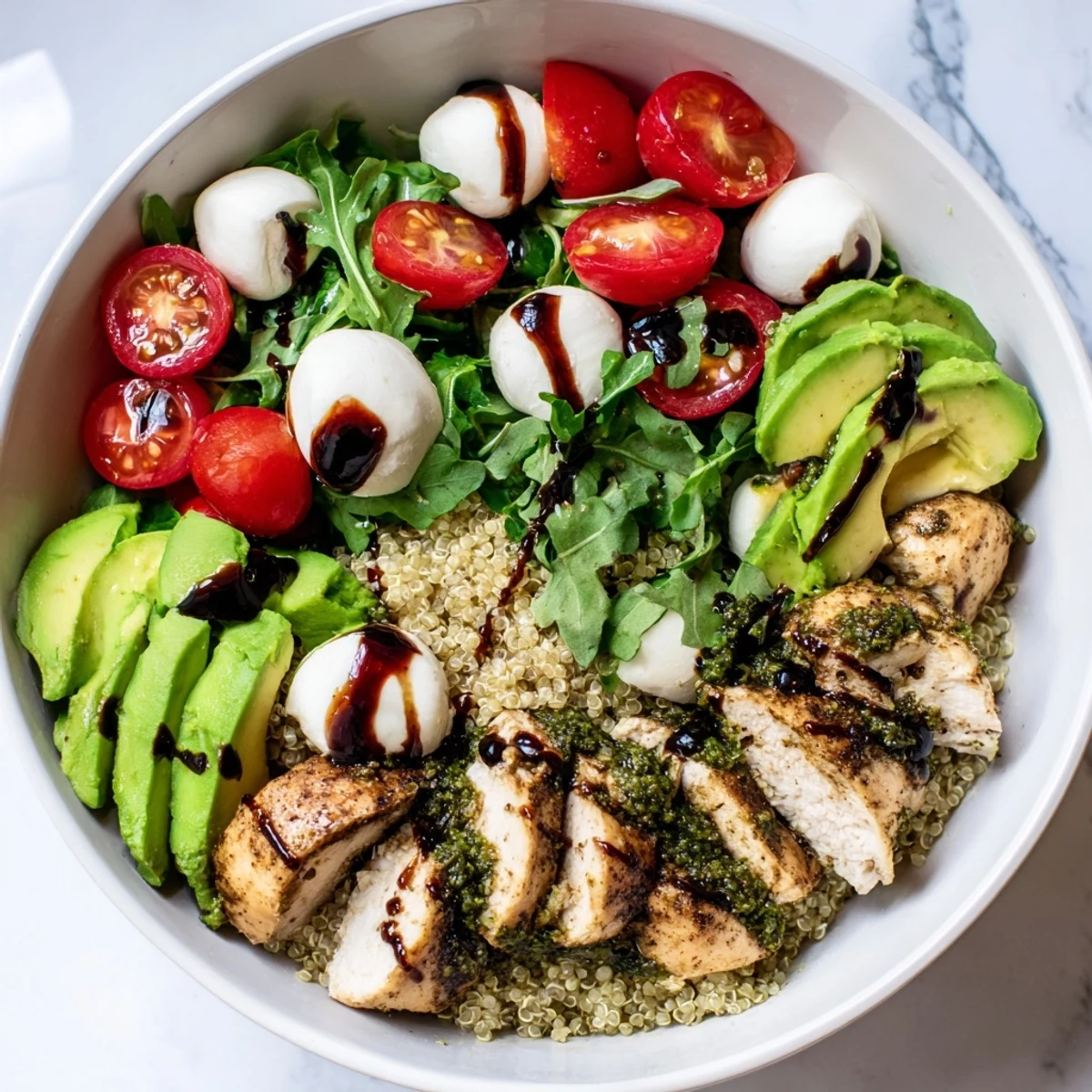 Close-up of a vibrant Pesto Chicken Caprese Grain Bowl featuring juicy pesto-marinated chicken, fresh mozzarella, halved cherry tomatoes, sliced avocado, and torn basil leaves on a bed of fluffy quinoa, all drizzled with balsamic glaze.